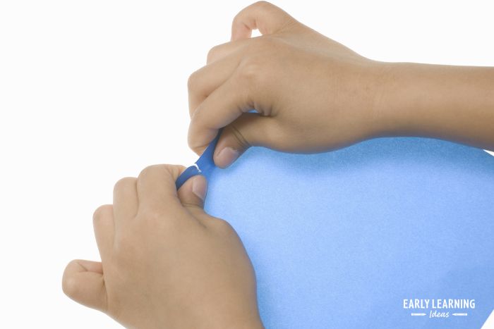 A child tearing paper for a fine motor activity.  The child is pinching a piece of blue paper with both hands to rip it into a strip for tearing paper activities.