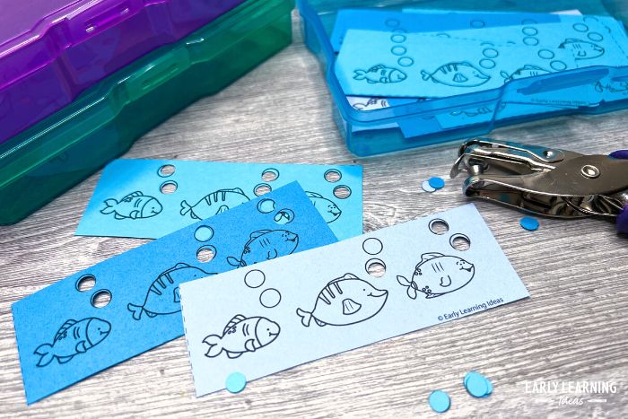 a free hole punch activity printed on different colors of blue paper. The fish themed activities with hole punched bubbles are pictured with a blue photo storage box and a hole punch..  You will love this cute and free hole punch printable for kids