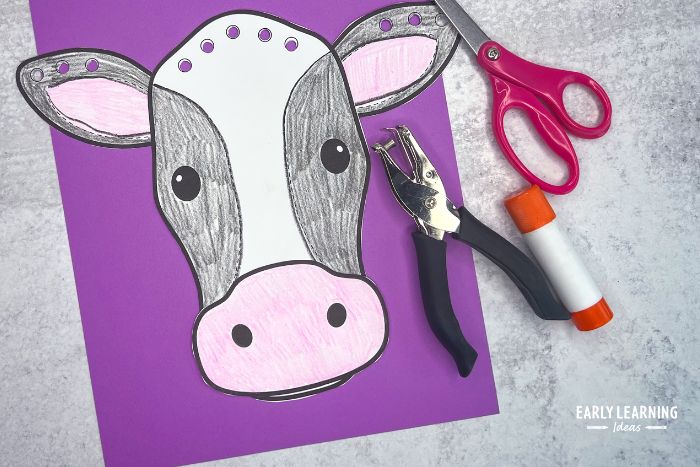 A black and white cow craft made of paper with a scissors, hole punch, and glue stick as an example of farm crafts for preschoolers.
