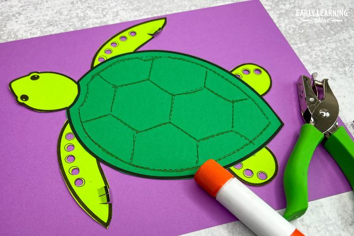 A sea turtle craft activity printed on two color of green paper.  The sea turtle craft is pasted to a purple background and is shown with a glue stick and a hole punch. - an example of ocean crafts for preschool.
