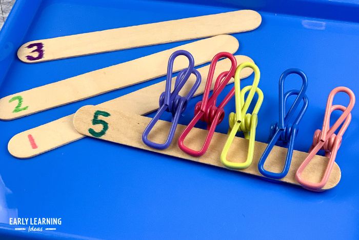 5 metal clothesline clips or chip clips on a craft stick with the number 5 written on it.  This is an example of dollar tree preschool activities and ideas to help kids build fine motor skills with inexpensive supplies.
