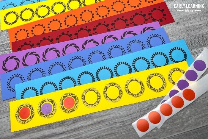 A bunch of colorful, printable dot sticker activity strips. A yellow strip is in front with some red and purple stickers. The stickers have been applied in an abab pattern. An example of dot sticker activities to build fine motor skills. -an example of dot sticker printables used for simple fine motor activities