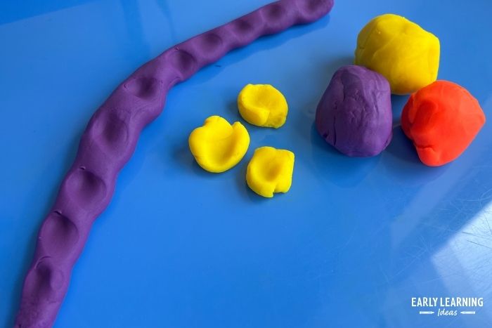 purple, yellow, and orange playdough on a blue tray.  A child has rolled a coil of purple playdough and pinched it along the coil - an example of pincer grasp activities.