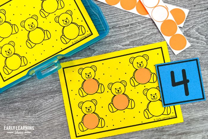 Teddy bear fine motor task cards printed on bright yellow paper.  A card has 5 bears on it, 4 of the bears have a dot sticker on them, and it is shown with a number 4 card.
