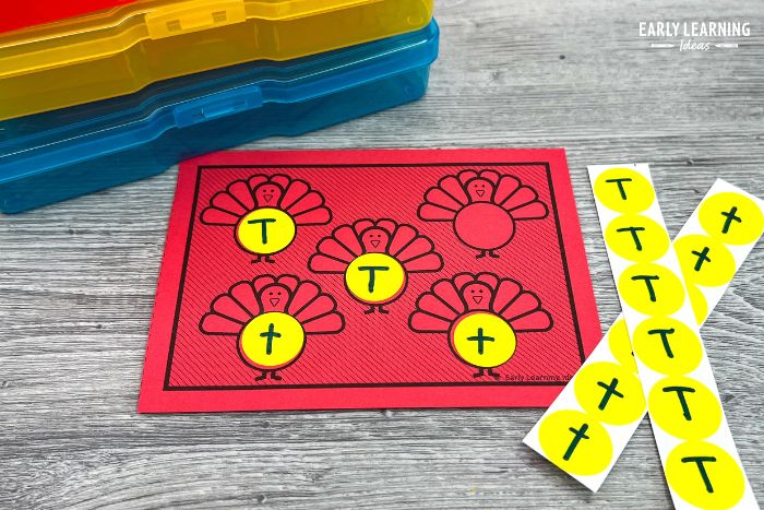 A turkey them fine motor task card printed on bright red paper and show with some plastic task boxes and dot stickers with the letter t written on them. - an example of a fine motor task card and letter sound activity for preschoolers.