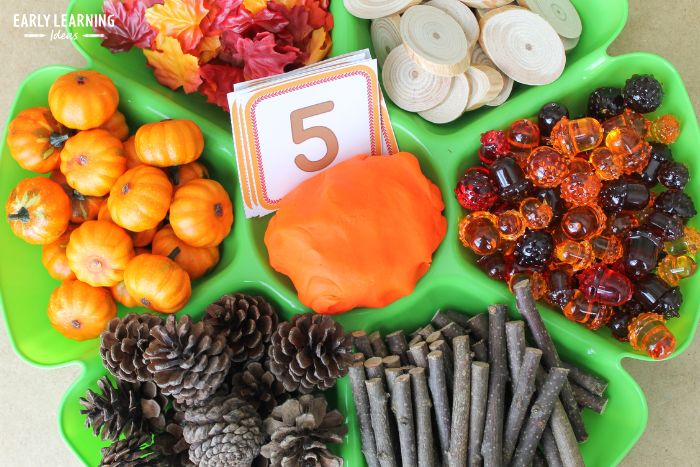 playdough activities for fall - a playdough tray with orange playdough, pumpkins, pine cones, sticks, plastic acorns, wood cookies, and silk fall leaves.