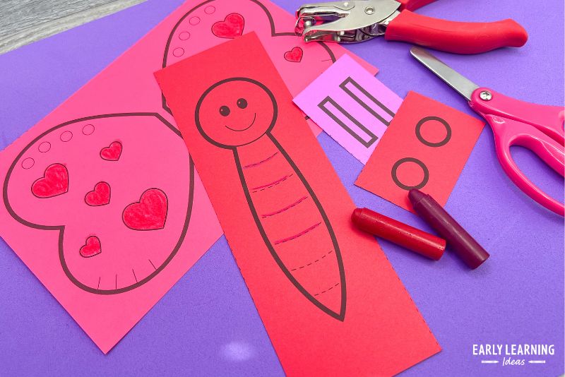 A love bug Valentine's Day 
craft template printed on red and pink paper shown with crayons, scissors, and a hole punch.
