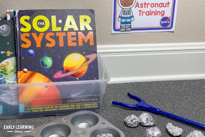 A box of non-fiction space books, pretend space rocks, and a toy grabber in a space theme dramatic play area