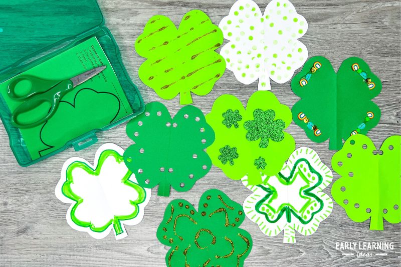 A variety of shamrock cutouts decorated with glitter glue, stickers, markers, etc.  It is shown with a task box of printable templates and a scissors - and example of St. Patricks Day crafts for preschool kids