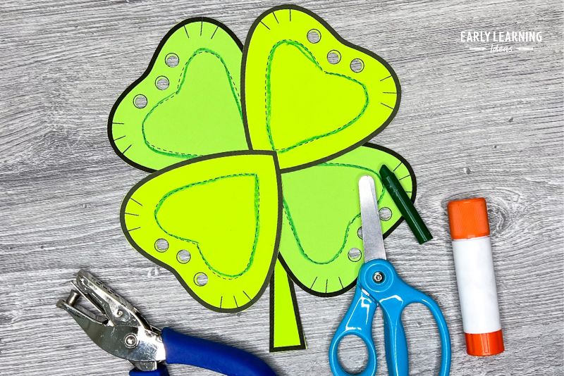 A shamrock cut and paste craft shown with scissors, a crayon, a glue stick, and a hole punch.  An example of St Patricks Day crafts for preschool kids.
