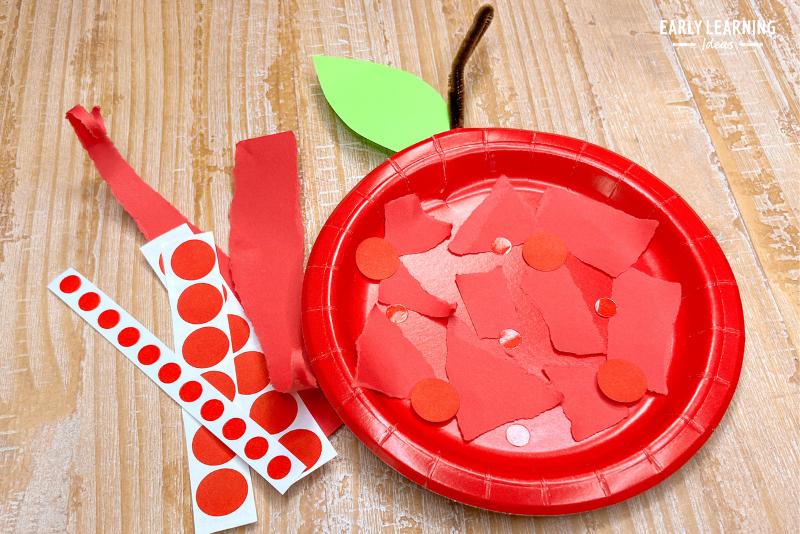 A paper plate apple craft. Red paper and dot stickers were added to the paper plate. A green construction paper leaf and a brown pipe cleaner stem complete the craft