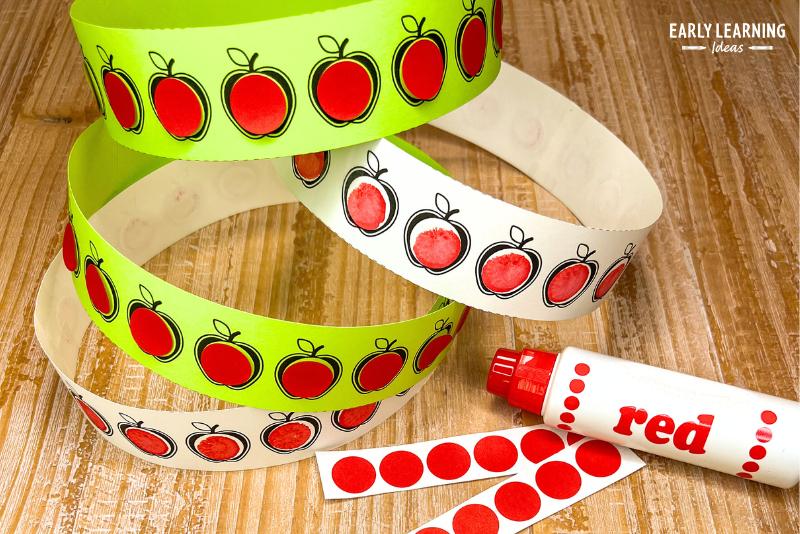 An apple headband made with apple strips printed on green and white paper. The apples are filled with red dot stickers or a dot from a bingo marker.
