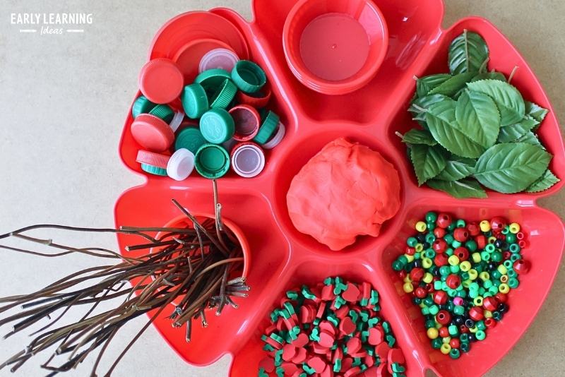 an apple themed playdough tray filled with red playdough, bottle caps, beads, leaves, sticks, and apple erasers