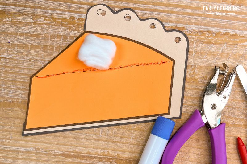 Paper pumpkin pie craft with cotton ball whipped cream shown with glue stick , hole punch, scissors