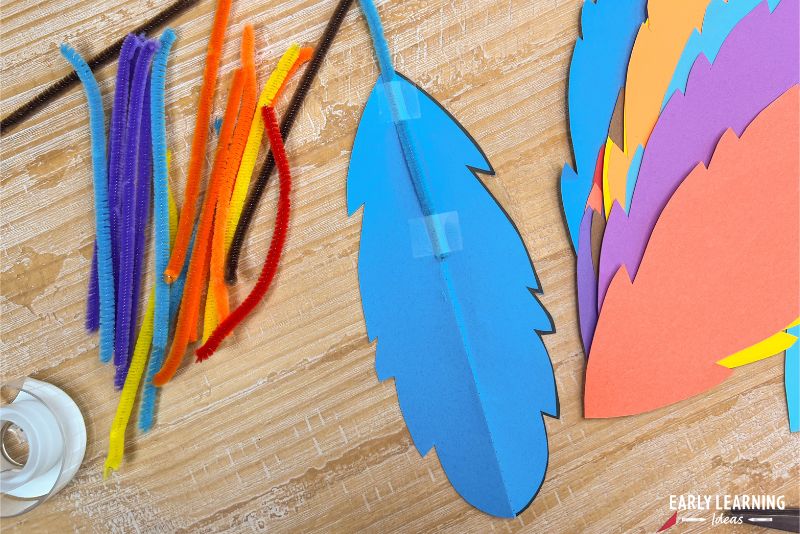 attaching a pipe cleaner to a blue paper feather with tape