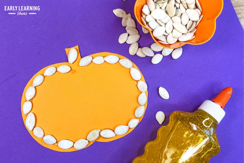 Pumpkin craft - an orange paper cutout is outlined with pumpkin seeds.  The craft is shown with a bowl of pumpkin seeds and a bottle of yellow glitter glue.