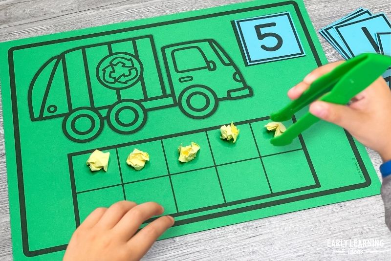 Earth Day counting mat with a recycling truck on it. A number 5 card is on the mat and a child is using large tweezers to place small balls of paper on the mat.