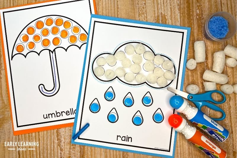 umbrella and rain cloud dot sticker crafts for an April showers theme shown with dot markers, crayons, scissors, and packing peanuts