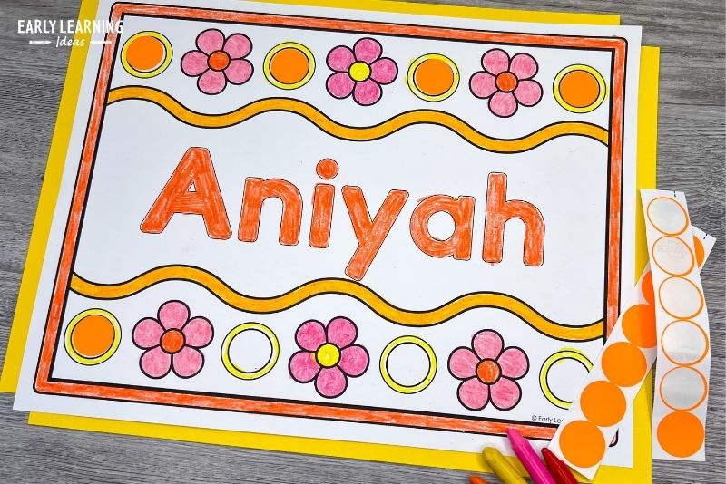 A flower name coloring page with the name Aniyah printed on it.  The page is colored and circles on the border are being covered with orange dot stickers.