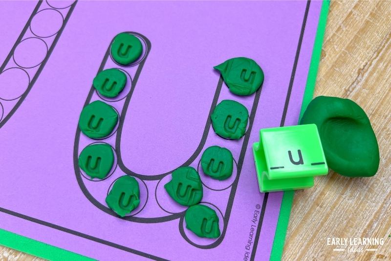 A letter u dot letter printable with small balls of green playdough in the dots. The playdough is then stamped with a lowercase letter u stamp so that a letter u is made in each circle.